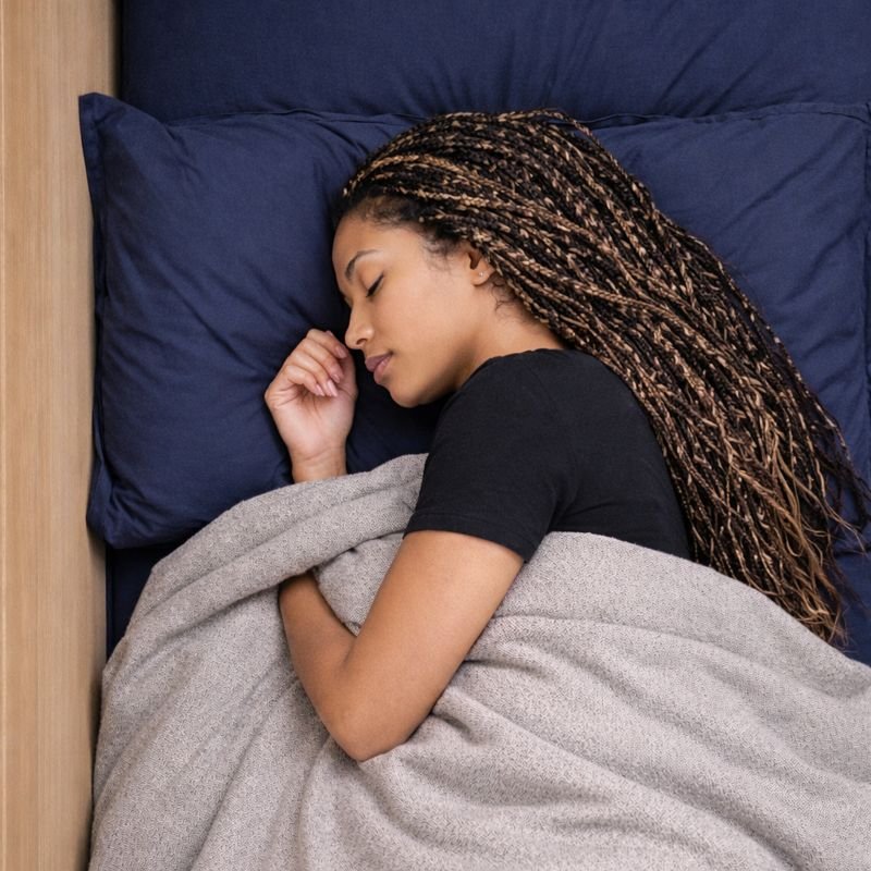 A person is peacefully sleeping in a bed, covered with a dark blue blanket. The bed is positioned near a wooden headboard, and the person is lying on a matching blue pillow. They are wearing a light blue long-sleeve shirt, with one arm resting on their pillow and the other partly under the blanket.